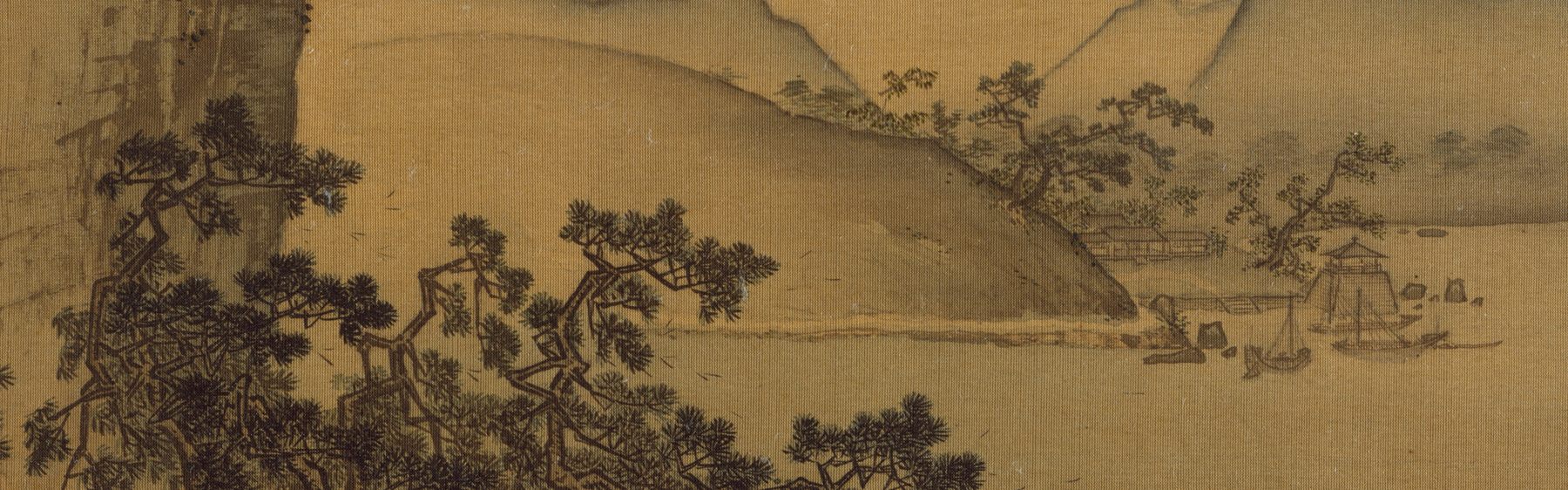 A wide ink landscape on aged golden silk: dark wind-bent pines crowd the foreground at the left below a cliff, a bare bluff rises behind them, and across calm water at the right small boats lie moored beside a thatched shelter, with range behind range of hills dissolving into mist.